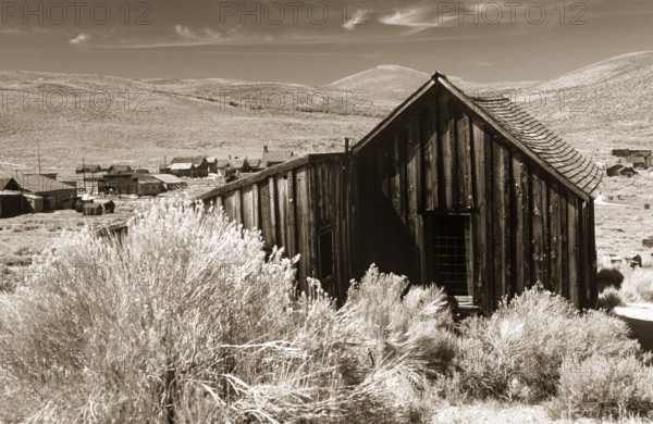 Old wooden shed in Bodie, behind it grassy hills of the prairie. Photo in sepia tone. Old wooden