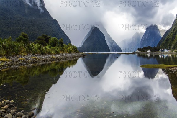 Mirror-smooth water of the Milford Sound fjord reflects mountains. Sunlight breaks through storm