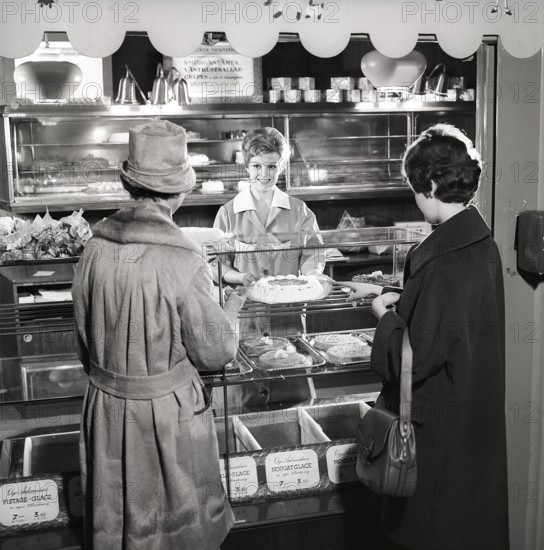 1960s coffee shop. Two women are standing in front of the counter that is full with freshly baked