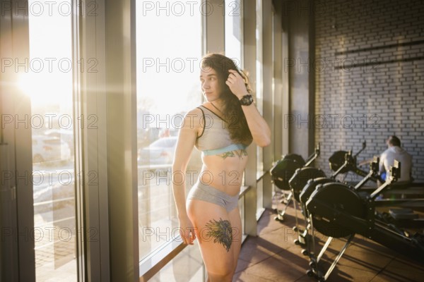 Young caucasian sporty woman with curly long hair poses in sportswear near a panorman window in a