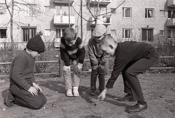 In the 1960s. Boys outdoors playing marbles. An old fashioned game where the goal is to hit the