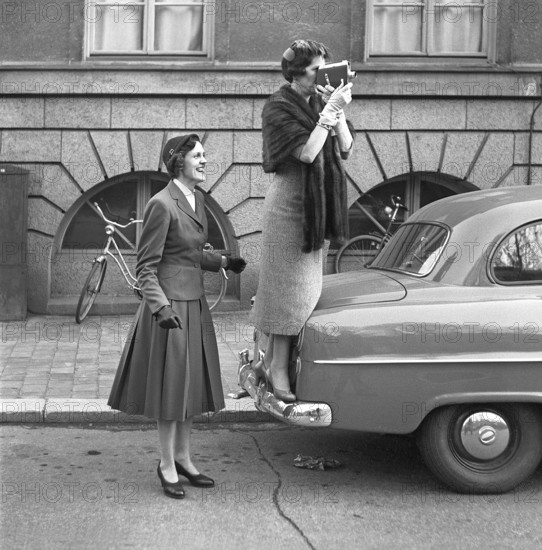 In the 1950s. An elderly woman with a film camera has stood on the bumper of a car to get a better