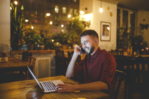 Young handsome Caucasian man with beard and toothy smile in red shirt works behind laptop, hands on