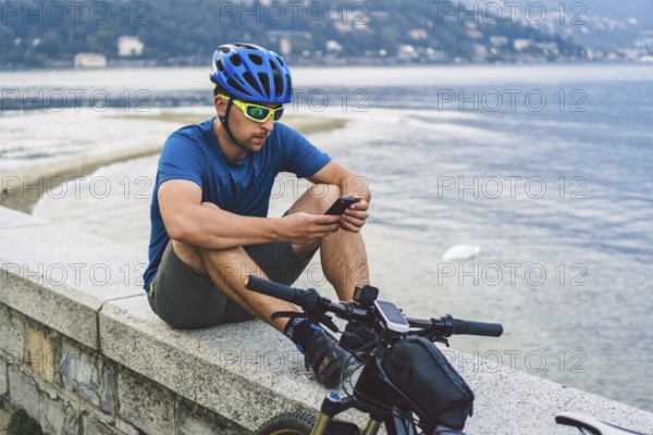The theme of tourism and travel in Italy. A male cyclist uses a phone on the shore of Lake Como.