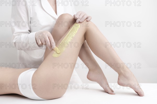 Cosmetologist applying green hot wax on slim woman leg using spatula while woman lying down on