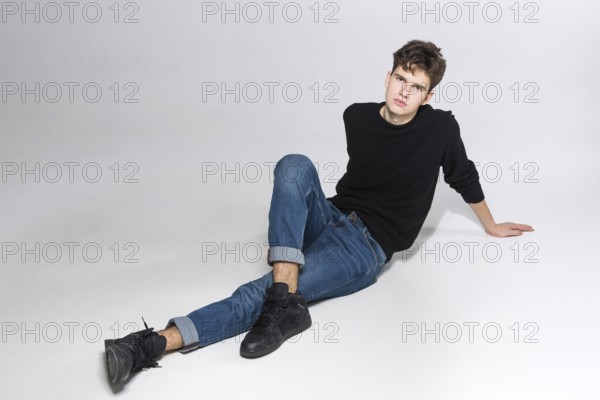 Stylish guy posing in studio sitting on the floor in denim pants and black sweatshirt. Male student