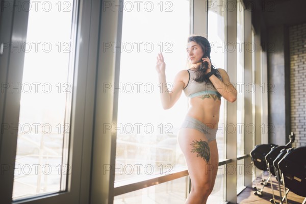 Young caucasian sporty woman with curly long hair poses in sportswear near a panorman window in a