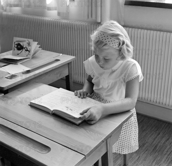 School in the 1950s. A girl is seen sitting at her desk in the classroom reading from the