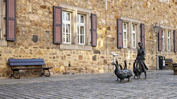 Scene in front of a stone building with shutters, a bench and a statue of a girl with geese, The
