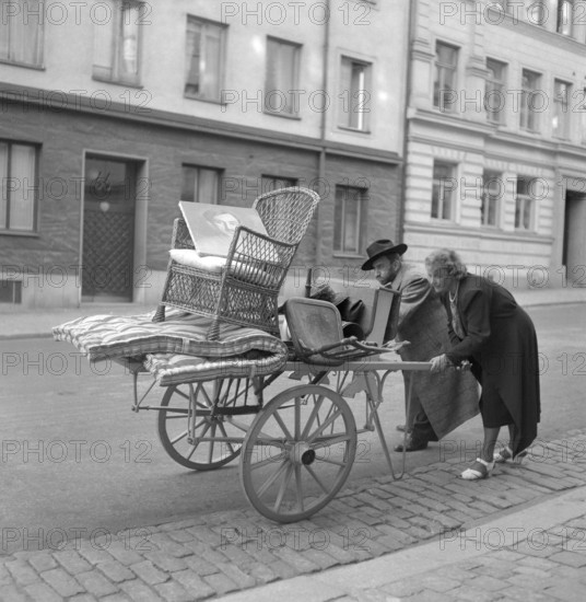Moving in the 1940s. A couple are pushing a carriage in front of them with some furniture on it.