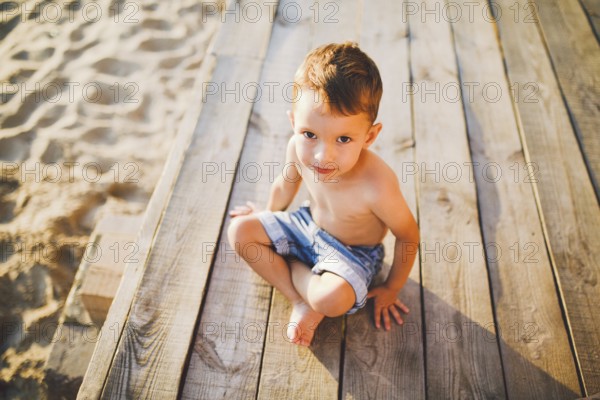 Little Caucasian boy child sitting on wooden pier sandy beach, summer time, sea holiday near water.