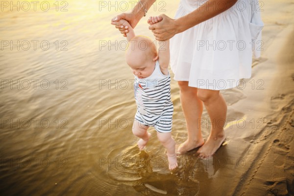 Baby boy walking along beach with mom's support. Mother teaching baby to walk. Outdoor, trust