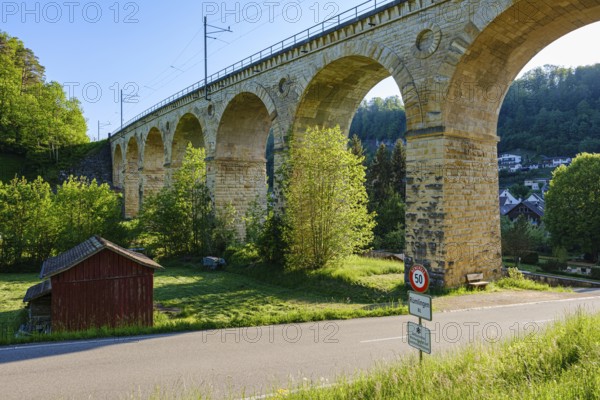 A massive stone viaduct towers over a green landscape with a small country road in the foreground,