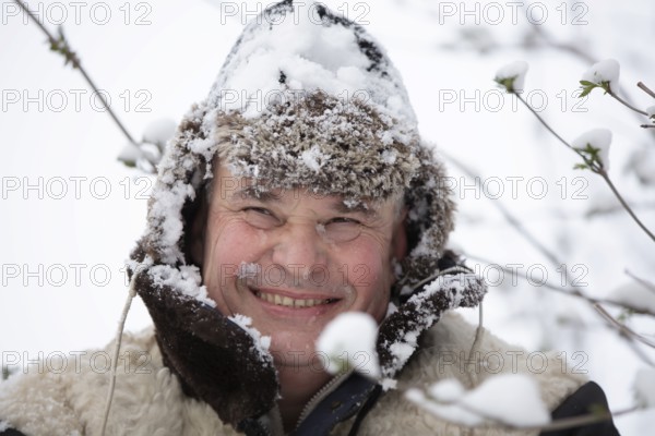 An elderly man in a snowy winter hat smiles and looks into the camera