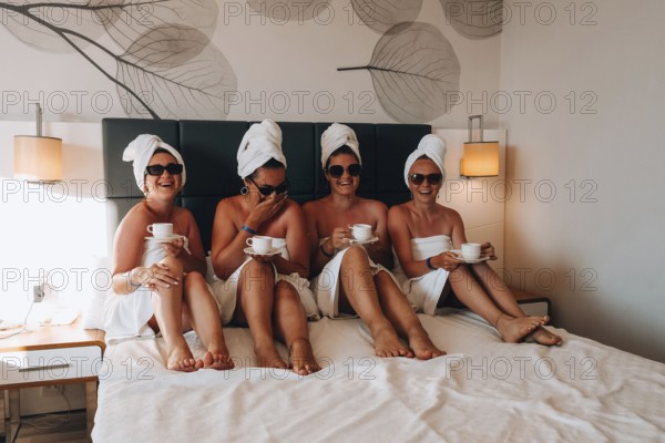 Four girls friends in white towels sitting on the bed having fun in hotel. Vacation Bloggers