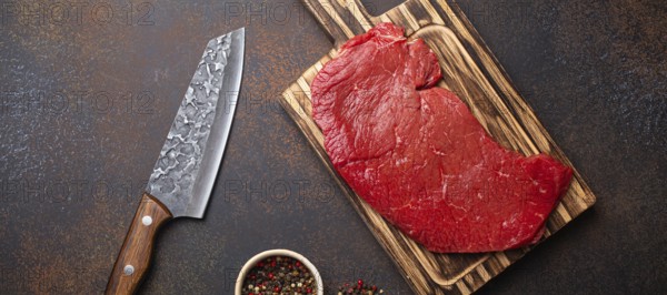 Raw uncooked top round beef steak on wooden cutting board with big kitchen knife and pepper on dark