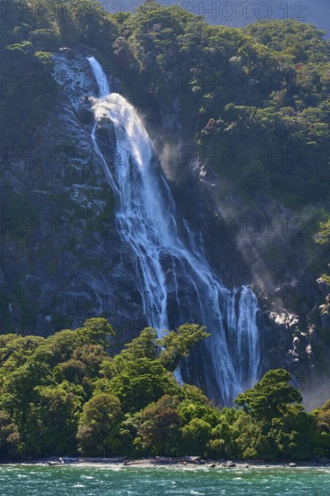 Waterfall cascades over a wooded slope and sprays fine spray into turquoise-coloured water, summer,
