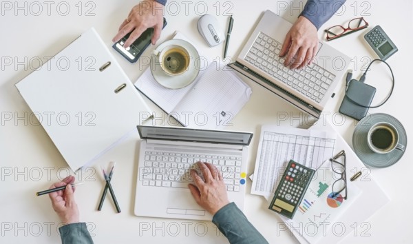 Top view of a white office desk with hands of two people working on laptops with papers,