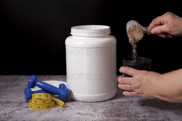 Woman pouring protein powder into a shaker with a spoon, next to a bottle of protein, dumbbells and