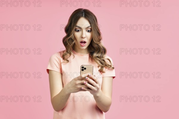 Portrait of a shocked pretty millennial hipster girl using mobile phone on pink background.