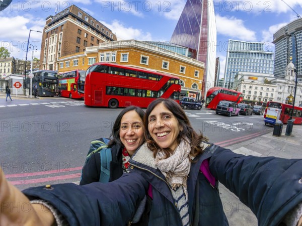 Two happy female tourists taking a selfie with red double decker buses and modern skyscrapers in
