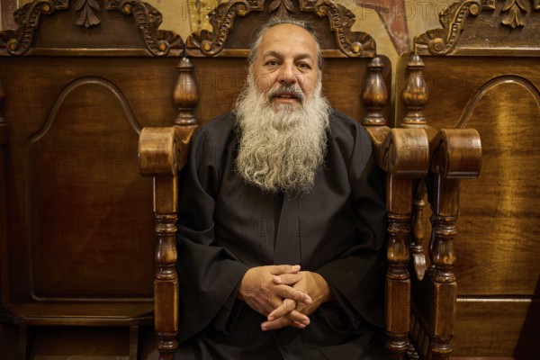 Papas Nektarios, Elderly man with beard on a wooden chair in a quiet, monastic environment,
