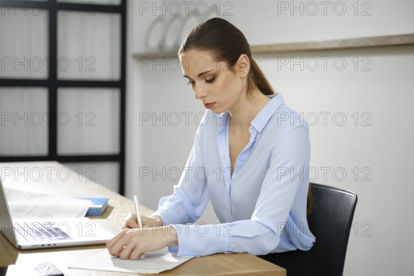 A business woman sits at a wooden desk, intently writing on a piece of paper while using her laptop