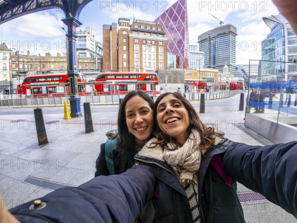 Two happy tourists taking a selfie in the city of london, with red buses and modern buildings in