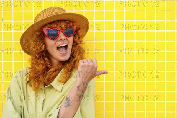 Excited young woman with red hair, heart shaped sunglasses, and straw hat pointing her thumb to the