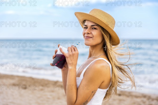 Young woman in a straw hat enjoying a refreshing drink while relaxing on the beach, soaking up the