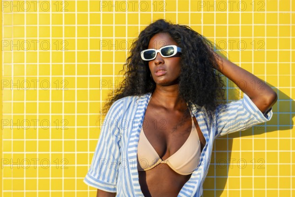 Stylish model with sunglasses and striped shirt posing against a vibrant yellow tiled wall,