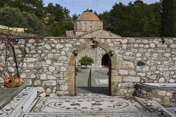 Path of mosaic stones leads to a church, surrounded by a high stone wall and trees, Thari