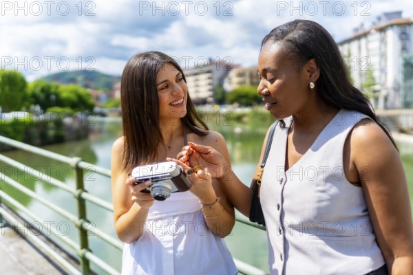 Two female friends looking at a vintage camera, standing next to a river in a sunny city, enjoying