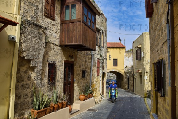 Narrow historic alley with a scooter driver and flower pots on a stone building, Rhodes Old Town,