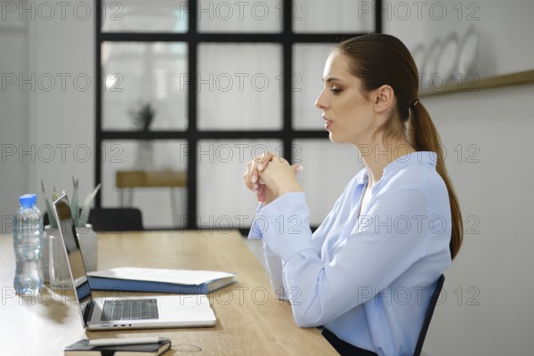 A woman wearing a light blue blouse sits at a modern desk engaged in a virtual meeting,