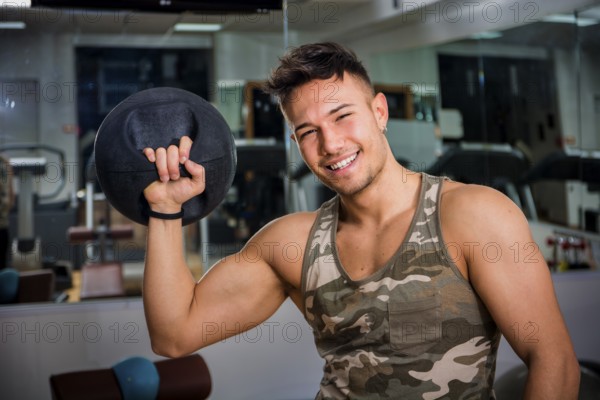 Attractive young athletic man exercising in gym, working out using kettlebell, looking at camera