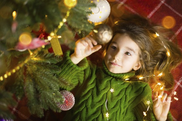A girl with long hair and garlands lies on a red plaid under a Christmas tree with toys in a warm