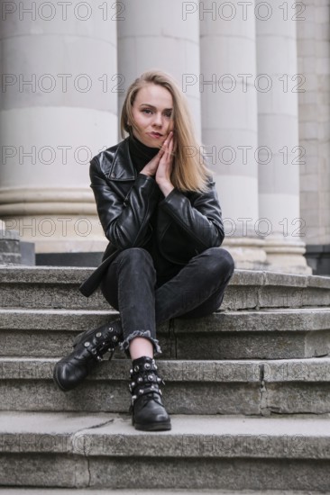 Portrait of young blonde woman with red lips wearing leather jacket on corporate building stairs.