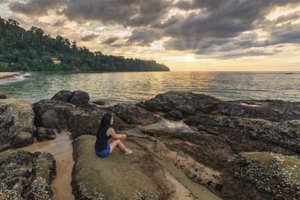 Tropical sea beach sunset with woman tourist on rock coast and ocean wave at Khao Lak, Phang Nga