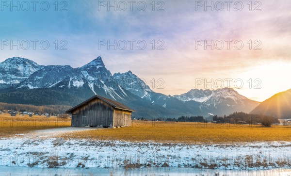 Winter alpine landscape with an aged wooden barn, the Alps mountains, a frozen river at sundown.