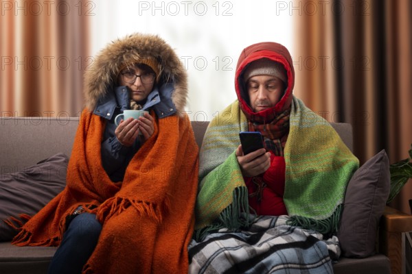 Middle age family in parkas drinking coffee on the sofa while using smartphone in their cold living