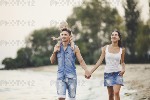 A young family of three walks along a sandy beach. A little baby girl with a blonde one year old