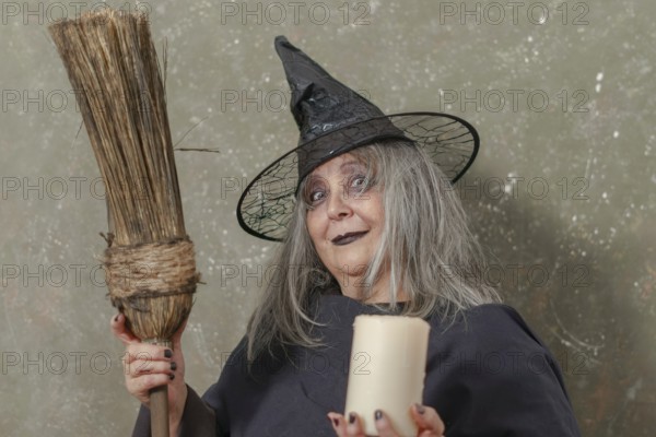 Old witch with white hair and a black hat holding a broom in her hand and a candle in the other