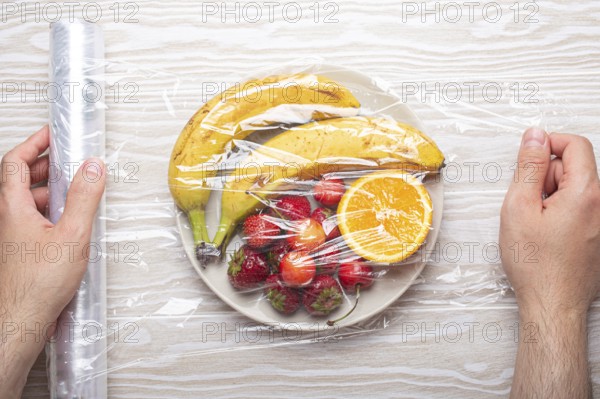 Male hands wrapping assorted different fruit and berries on white plate in plastic clear film top