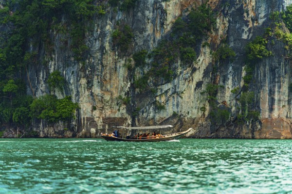 Longboat navigating along a towering cliff with greenery above turquoise water, Cheow Lan Lake,