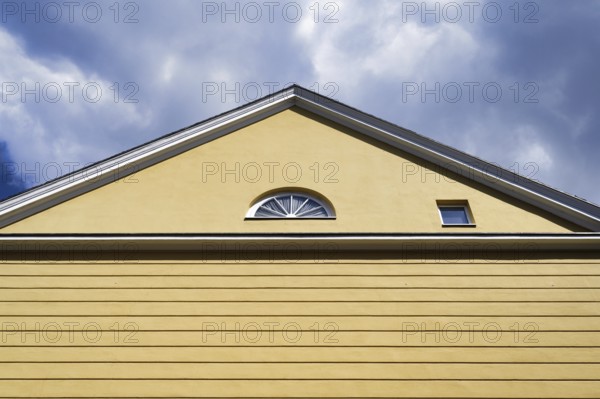 A yellow house with a pitched roof and one semi-circular and one square window in front of a cloudy