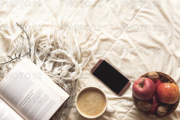 Coziness concept with an open book on a wool shawl, a basket with apples and nuts, a phone and a