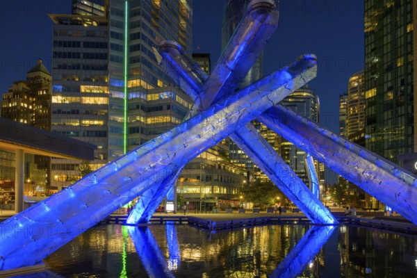 Modern Olympic fire installation at night with blue lighting and surrounding skyscrapers in the