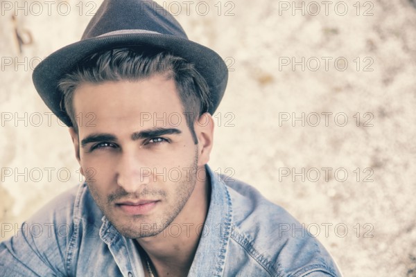 Handsome young, man in denim shirt and fedora hat sitting in pose on sandy beach and looking at