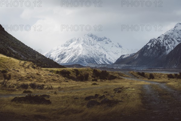 Gravel track leads through a vast landscape with snow-capped mountains and cloudy skies, Hooker